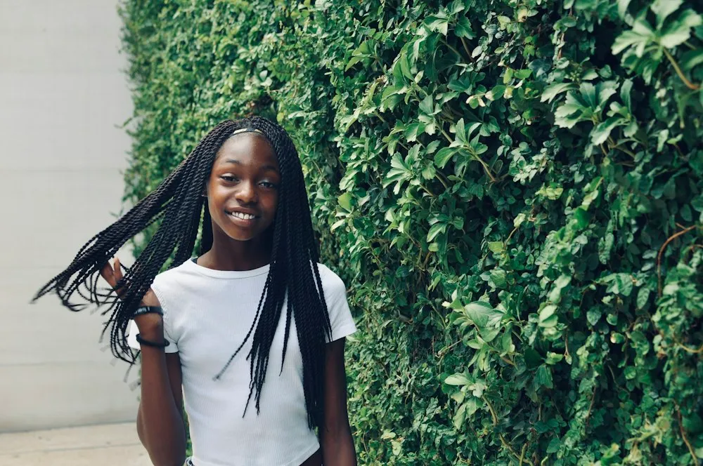 A teenage girl with long braids standing by a green hedge