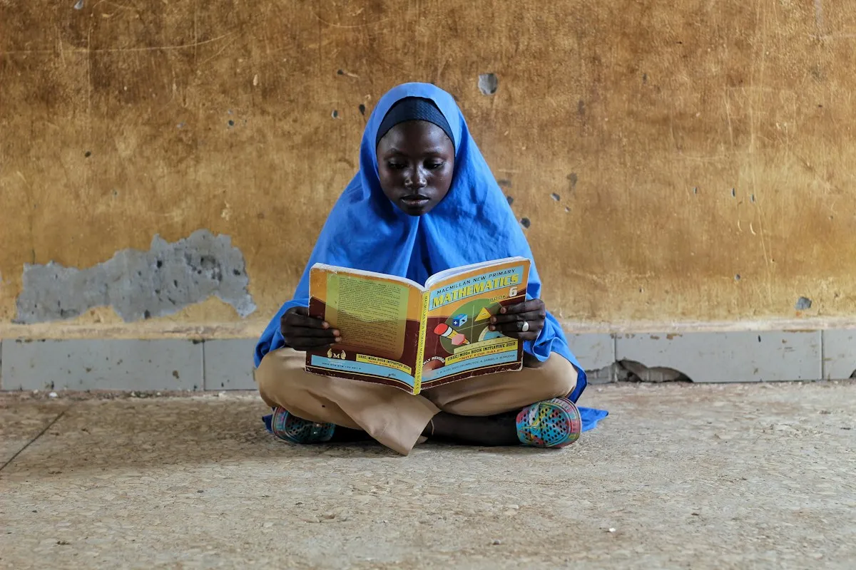 A schoolgirl in a blue headscarf sitting cross-legged, reading a book